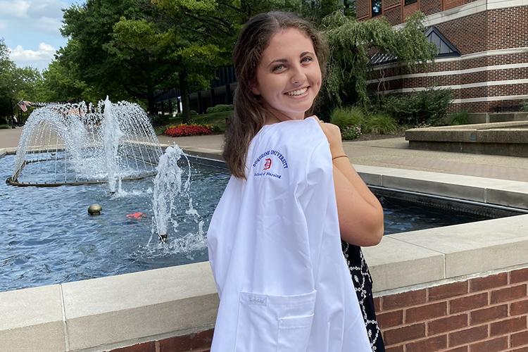Young woman, poses with white coat over shoulder and smiles at camera, in front of water fountain